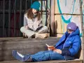 Dwoje dzieci czyta na schodkach. Dziewczynka w turkusowej czapce pochyla się nad książką, a chłopiec w fioletowej czapce przegląda lektury obowiązkowe.