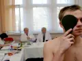 Młody mężczyzna zasłania oko podczas badania wzroku. W tle lekarze i urzędnicy pracują, ustalając, ile trwa komisja wojskowa.