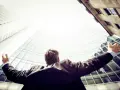 Mężczyzna w garniturze, z rękami rozłożonymi na boki, patrzy w górę na nowoczesne budynki. To symbol sukcesu, jakby pytał: "ceo co to znaczy?".