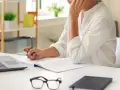 Osoba w białej koszuli pracuje przy biurku z laptopem, notatnikiem i okularami.