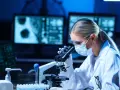 Diagnosta laboratoryjny w okularach i masce analizuje próbki pod mikroskopem. Na ekranach komputera widać wyniki badań.