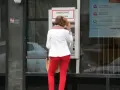 Kobieta w czerwonych spodniach i białej marynarce korzysta z bankomatu. Może to być symboliczne przedstawienie średnich zarobków w Polsce, gdzie ludzie polegają na bankomatach.