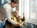 Mężczyzna w średnim wieku podaje starszemu mężczyźnie w wózku inwalidzkim tabletkę i szklankę wody.