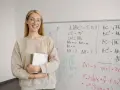 Uśmiechnięta nauczycielka z zeszytem przy tablicy z matematycznymi wzorami. Zastanawia się, ile punktów na maturze z matematyki można zdobyć za takie zadania.