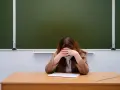 Zmęczony nauczyciel początkujący w maseczce siedzi przy biurku, chowając twarz w dłoniach. Przed nim leży zeszyt.