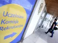 Znak "Uczelniana Komisja Rekrutacyjna" wskazuje drogę do budynku, gdzie trwa rekrutacja na studia. Mężczyzna z torbą idzie w kierunku drzwi.
