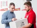 Kurier w czerwonej koszulce przekazuje paczkę, a klient podpisuje dokument. Zastanawiasz się, ile zarabia kurier?