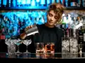 Młody barman nalewa drinka do szklanki. W tle widać półki z butelkami i niebieskie światła.