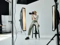 Młody fotograf z blond włosami, siedząc na stołku, robi zdjęcie aparatem. W studiu widać lampy i sprzęt. Zastanawiasz się, ile zarabia fotograf?