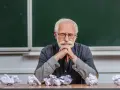 Zmęczony adiunkt z brodą i wąsami, w okularach, siedzi przy biurku z pogiętymi kartkami.