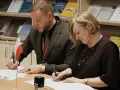 Para podpisująca dokumenty, z polską flagą obok. W tle półki z książkami, w tym te dotyczące slawistyki.