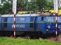 Niebieska lokomotywa PKP Cargo ET22-989 na tle zieleni i budynków.
