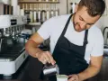 Uśmiechnięty barista w czarnym fartuchu nalewa mleko do kawy. Obok stoi profesjonalna ekspres do kawy.