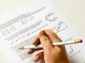 Ręka z ołówkiem rozwiązuje test fizyczny. Pytanie 22 dotyczy satelitów, a pytanie 23 ruchu po torze w kształcie podkowy. Od ilu procent jest 5?