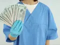 Osoba w niebieskim fartuchu i rękawiczkach trzyma wachlarz banknotów 20 dolarowych.