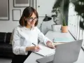 Uśmiechnięta redaktor w okularach notuje pomysły w zeszycie przy laptopie.