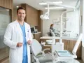 Uśmiechnięty dentysta w białym fartuchu trzyma narzędzia. W tle gabinet stomatologiczny, gdzie można dowiedzieć się, ile zarabia dentysta.