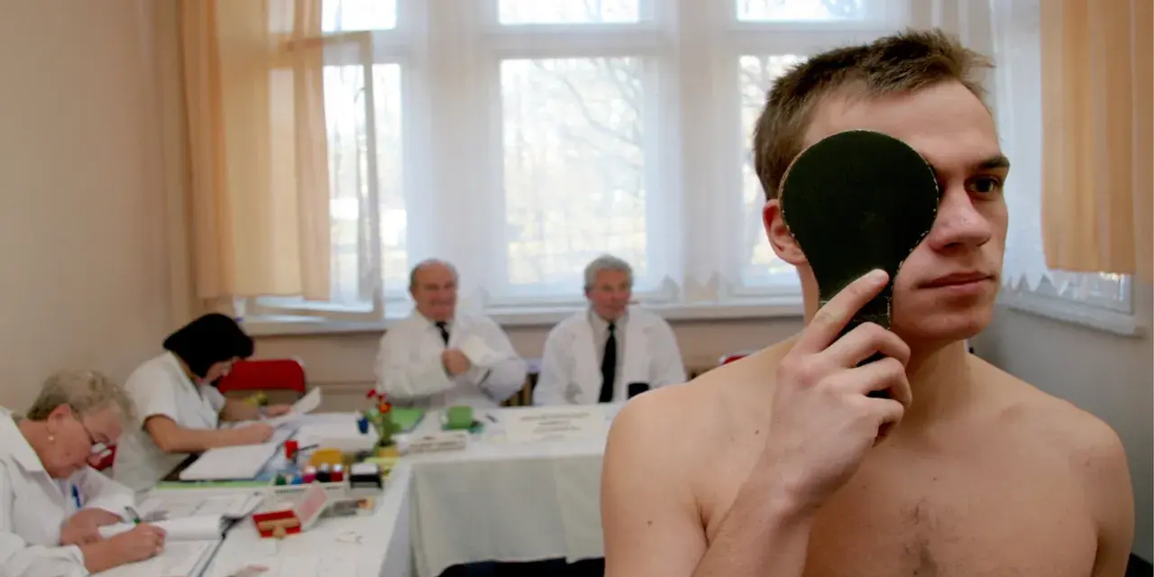 Młody mężczyzna zasłania oko podczas badania wzroku. W tle lekarze i urzędnicy pracują, ustalając, ile trwa komisja wojskowa.
