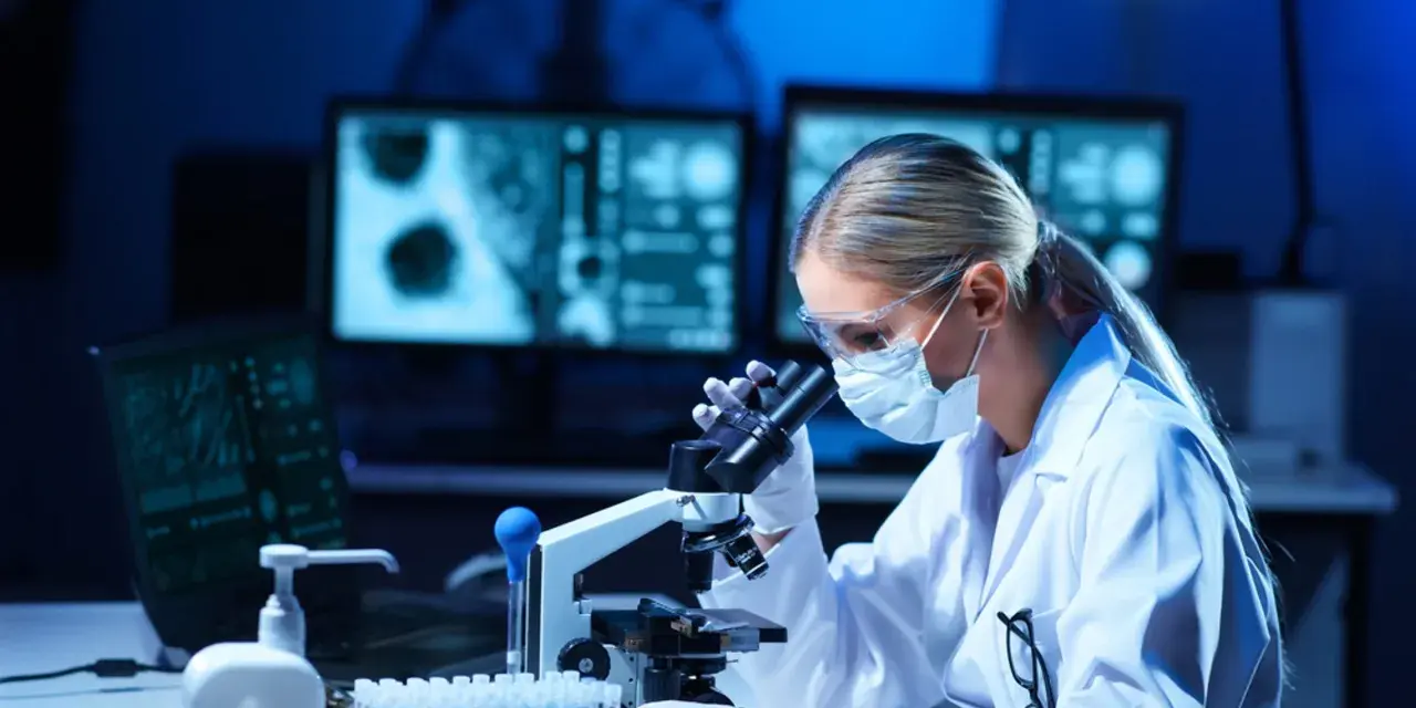 Diagnosta laboratoryjny w okularach i masce analizuje próbki pod mikroskopem. Na ekranach komputera widać wyniki badań.