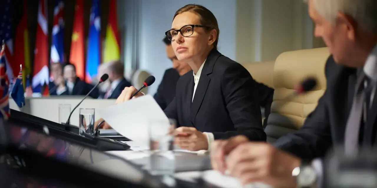 Kobieta w okularach analizuje dokumenty podczas konferencji międzynarodowej, gdzie omawiane są zagadnienia politologii.
