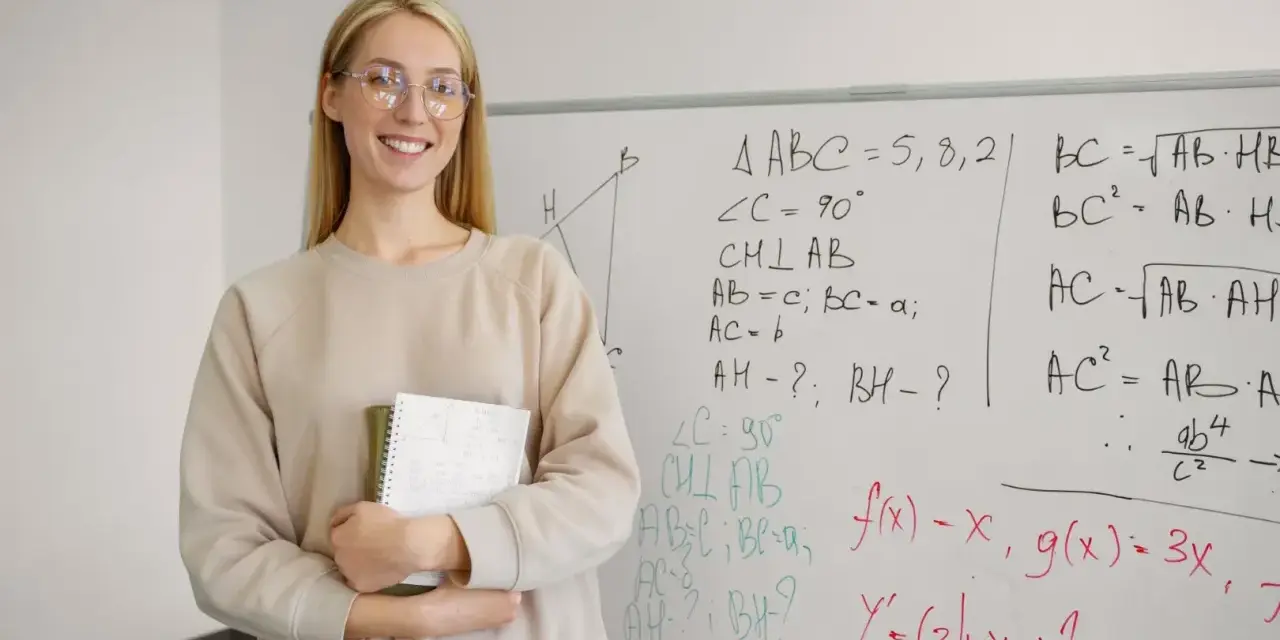 Uśmiechnięta nauczycielka z zeszytem przy tablicy z matematycznymi wzorami. Zastanawia się, ile punktów na maturze z matematyki można zdobyć za takie zadania.