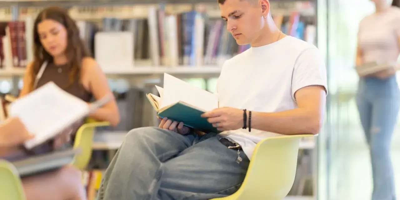 Młody mężczyzna w białej koszulce czyta książkę w bibliotece. W tle inne osoby i regały pełne książek, tworzące atmosferę nauki i spokoju, jakby czas przedwiośnia sprzyjał refleksji.