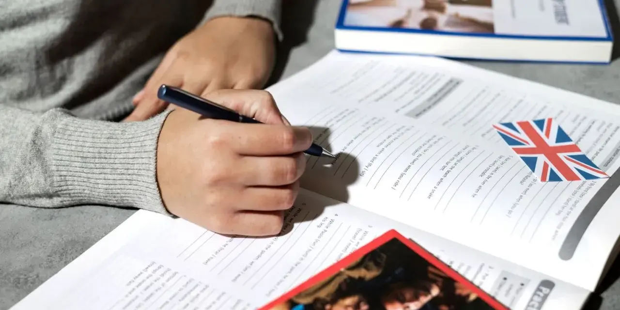 Osoba pisze długopisem w zeszycie z ćwiczeniami z języka angielskiego. Obok leży książka i flaga Wielkiej Brytanii. To pomoc w nauce, jak klauzula CV po angielsku.