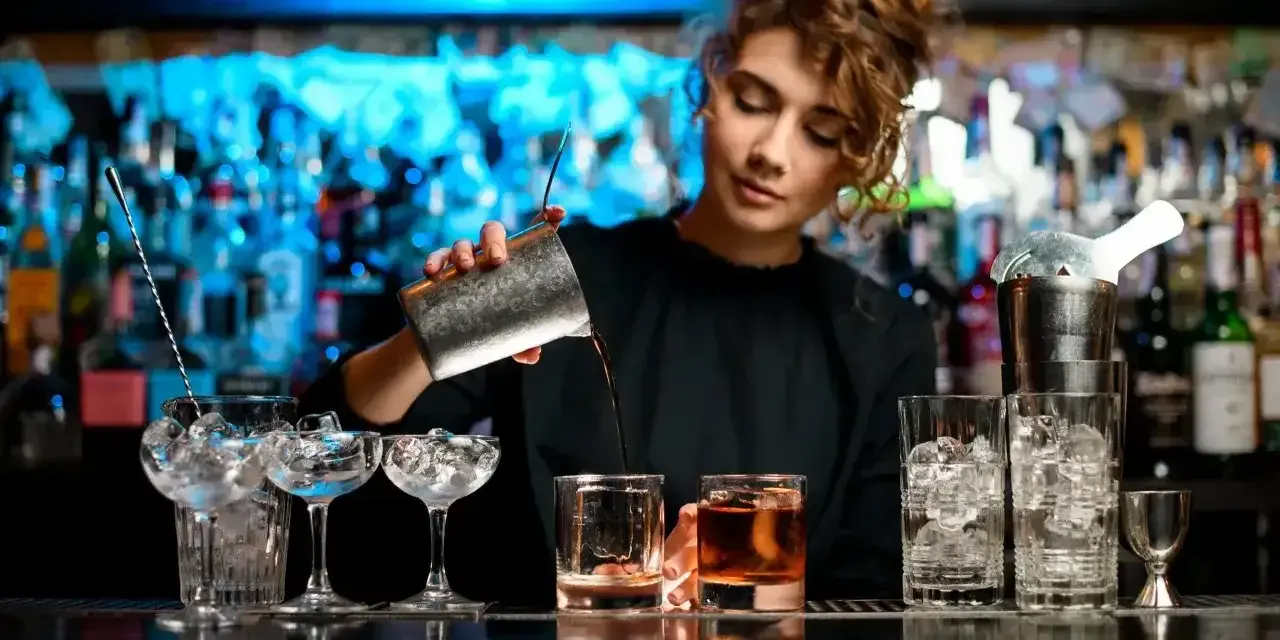 Młody barman nalewa drinka do szklanki. W tle widać półki z butelkami i niebieskie światła.