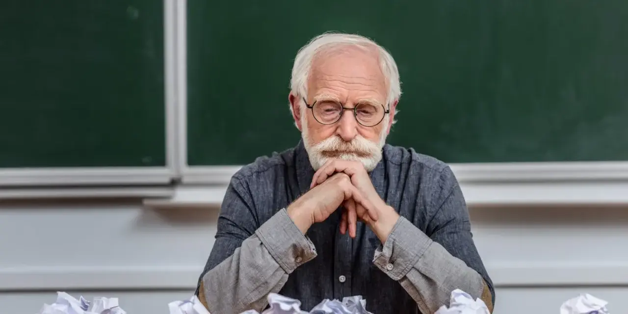 Zmęczony adiunkt z brodą i wąsami, w okularach, siedzi przy biurku z pogiętymi kartkami.