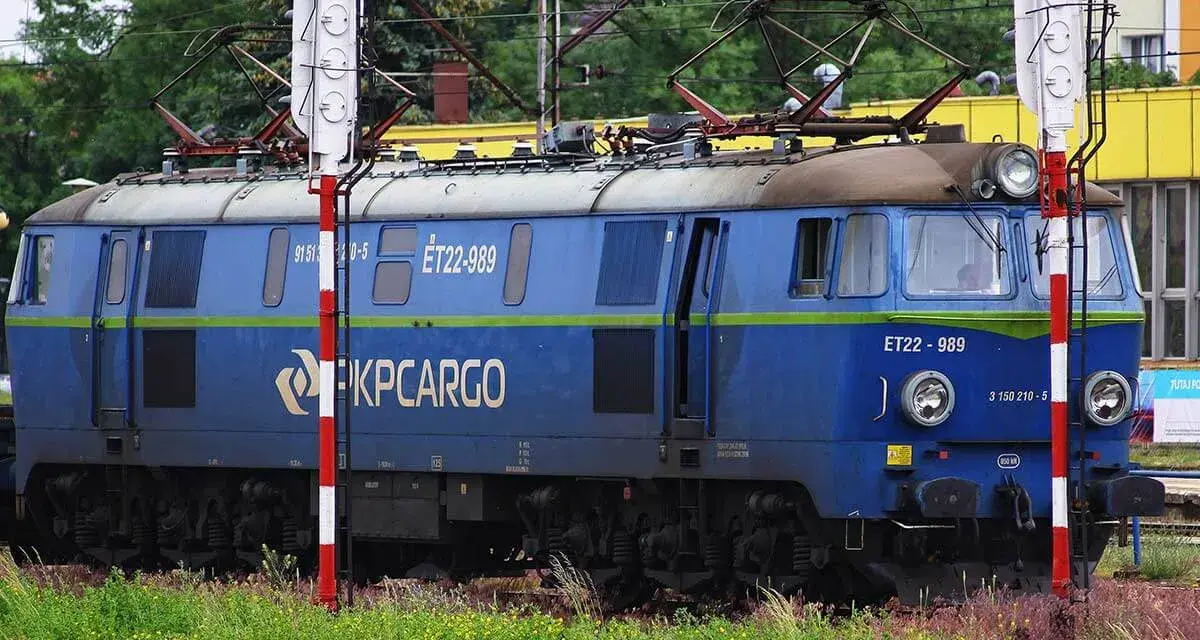 Niebieska lokomotywa PKP Cargo ET22-989 na tle zieleni i budynków.