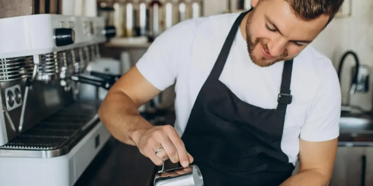 Uśmiechnięty barista w czarnym fartuchu nalewa mleko do kawy. Obok stoi profesjonalna ekspres do kawy.