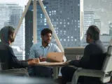 Trzech mężczyzn w biurze rozmawia przy stole. Jeden z nich, z laptopem, gestykuluje, jakby opowiadał coś coo.