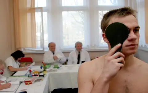 Młody mężczyzna zasłania oko podczas badania wzroku. W tle lekarze i urzędnicy pracują, ustalając, ile trwa komisja wojskowa.