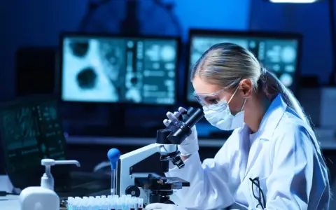 Diagnosta laboratoryjny w okularach i masce analizuje próbki pod mikroskopem. Na ekranach komputera widać wyniki badań.