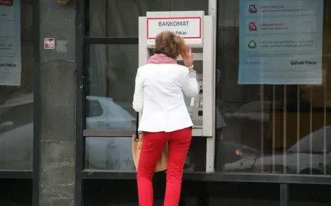 Kobieta w czerwonych spodniach i białej marynarce korzysta z bankomatu. Może to być symboliczne przedstawienie średnich zarobków w Polsce, gdzie ludzie polegają na bankomatach.