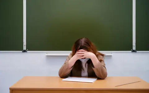 Zmęczony nauczyciel początkujący w maseczce siedzi przy biurku, chowając twarz w dłoniach. Przed nim leży zeszyt.