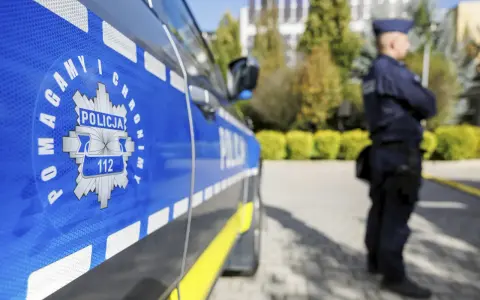 Policjant w mundurze stoi obok radiowozu. Czy zarobki w policji są adekwatne do służby?