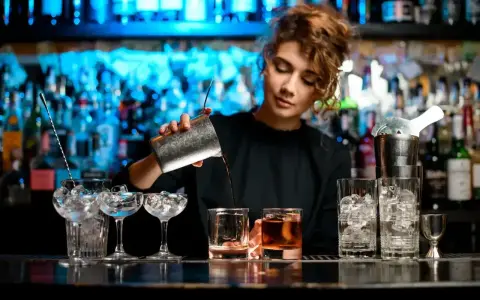 Młody barman nalewa drinka do szklanki. W tle widać półki z butelkami i niebieskie światła.