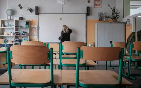 Nauczycielka przy tablicy, zastanawia się, ile zarabia przedszkolanka. Puste ławki w sali lekcyjnej.