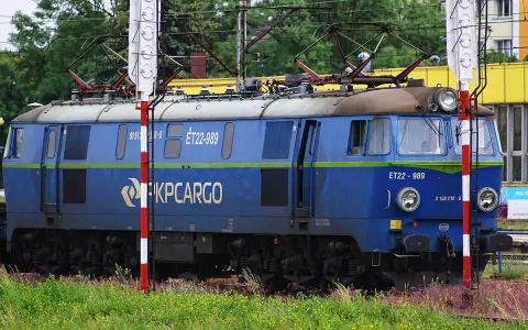 Niebieska lokomotywa PKP Cargo ET22-989 na tle zieleni i budynków.