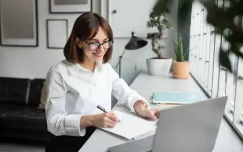 Uśmiechnięta redaktor w okularach notuje pomysły w zeszycie przy laptopie.