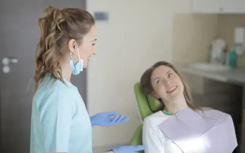 Uśmiechnięta pacjentka na fotelu stomatologicznym słucha dentystki. To obrazek z gabinetu, gdzie odbywają się studia stomatologiczne.