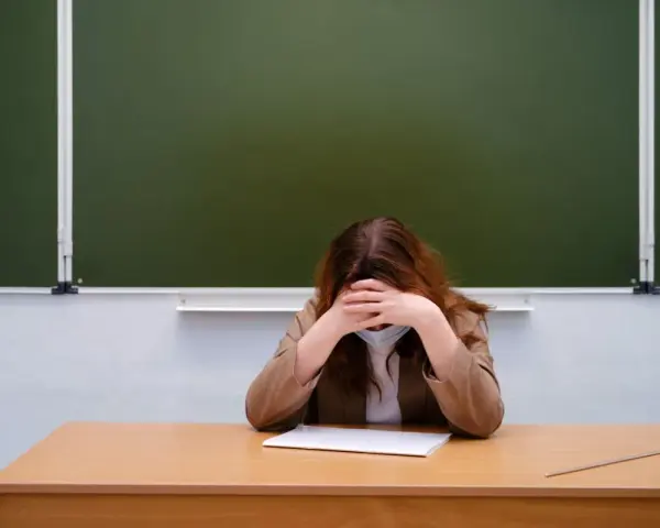 Zmęczony nauczyciel początkujący w maseczce siedzi przy biurku, chowając twarz w dłoniach. Przed nim leży zeszyt.