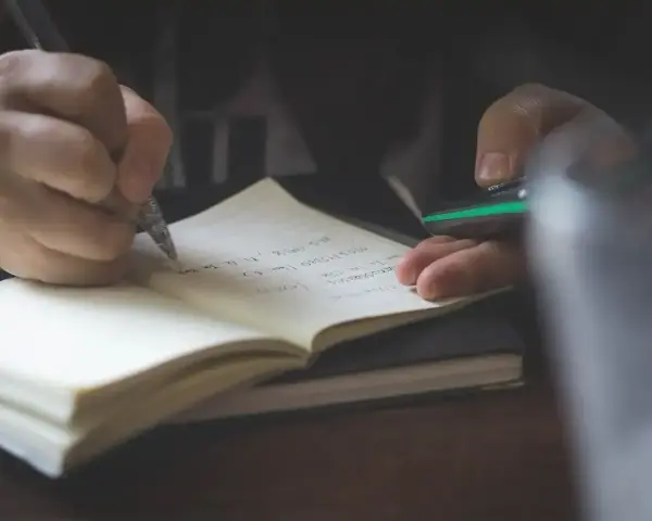 Dłoń pisze piórem w notatniku, przygotowując się do napisania rozprawki.