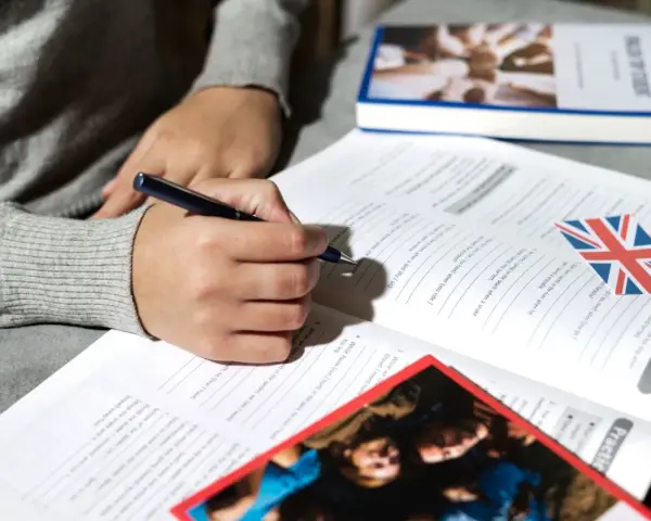 Osoba pisze długopisem w zeszycie z ćwiczeniami z języka angielskiego. Obok leży książka i flaga Wielkiej Brytanii. To pomoc w nauce, jak klauzula CV po angielsku.