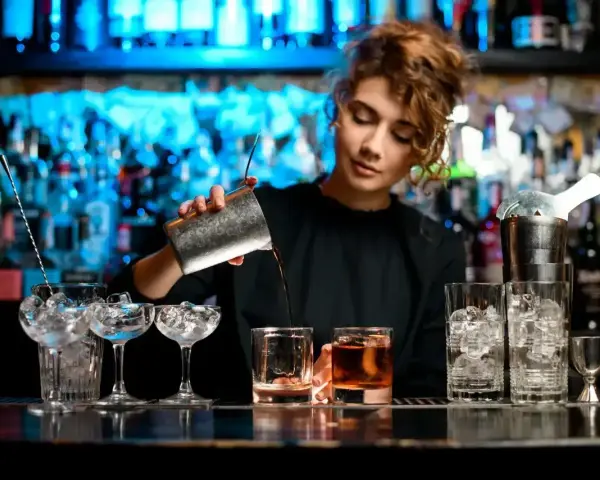 Młody barman nalewa drinka do szklanki. W tle widać półki z butelkami i niebieskie światła.
