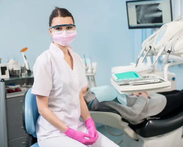 Asystentka stomatologiczna w masce i okularach ochronnych siedzi w gabinecie.