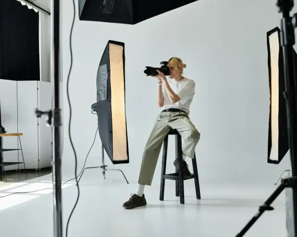 Młody fotograf z blond włosami, siedząc na stołku, robi zdjęcie aparatem. W studiu widać lampy i sprzęt. Zastanawiasz się, ile zarabia fotograf?