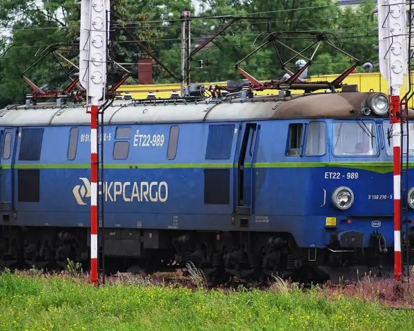 Niebieska lokomotywa PKP Cargo ET22-989 na tle zieleni i budynków.