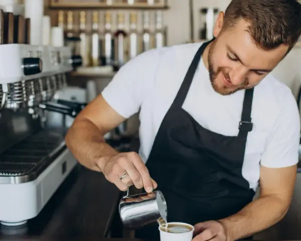 Uśmiechnięty barista w czarnym fartuchu nalewa mleko do kawy. Obok stoi profesjonalna ekspres do kawy.
